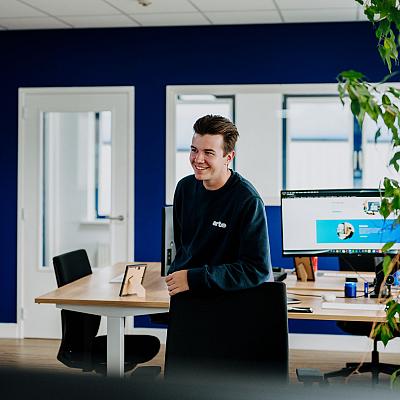 Een man in een donkere trui staat in een kantoor en kijkt naar een open laptop die hij in zijn handen houdt.
