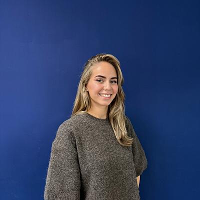 Een glimlachende vrouw met lang blond haar en een grijze top staat voor een blauwe wand.