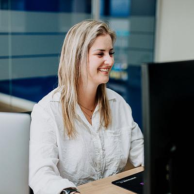 Vrouw met blond haar zit glimlachend achter een bureau en werkt aan een computer in een modern kantoor met glazen wanden.