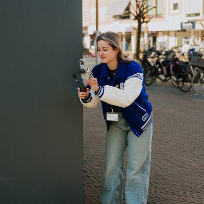 Jonge vrouw met blauwe jas filmt met een smartphone op een gimbal in een winkelstraat met fietsen en woonhuizen op de achtergrond.