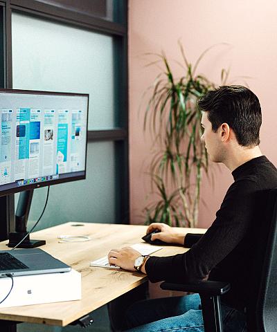 Man die achter zijn bureau werkt aan een grafisch ontwerp op een groot beeldscherm, in een modern kantoor met roze muur en plant op de achtergrond.