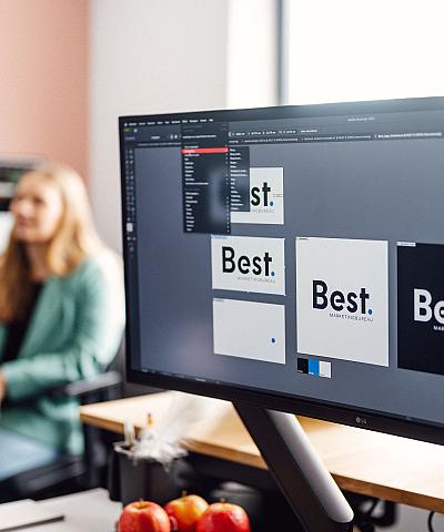 Beeldscherm waarop het logo van Best Marketingbureau wordt ontworpen in Adobe Illustrator, met op de achtergrond een vrouw in een turquoise blazer in een kantooromgeving.
