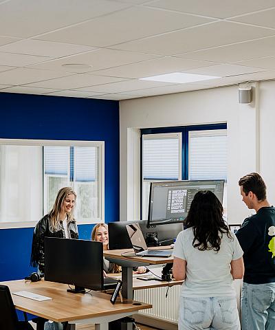 Vier collega’s staan en zitten samen rond een bureau in een modern kantoor met blauwe muren en grote ramen, terwijl ze iets bespreken op een computerscherm.