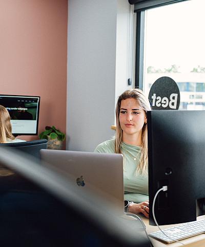 Vrouw werkt geconcentreerd achter twee computerschermen in een modern kantoor met roze muren en veel natuurlijk licht.