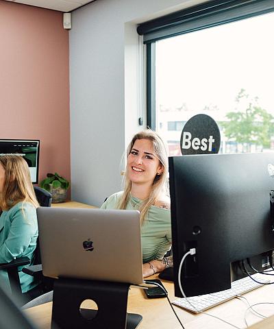 Twee collega’s die samen aan het werk zijn in een licht kantoor met grote ramen, waarbij een van hen lachend naar de camera kijkt.