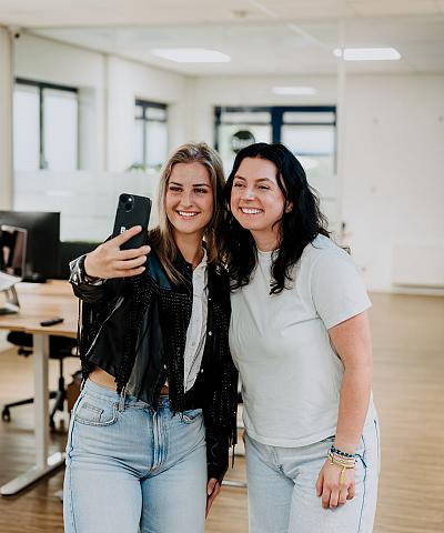 Twee vrouwen maken samen een selfie in een moderne kantoorruimte. Ze lachen en lijken plezier te hebben, terwijl ze voor de camera poseren.