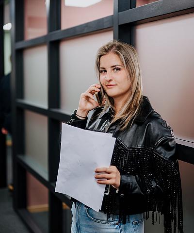 Medewerkster staat in een kantooromgeving terwijl ze telefoneert en een document vasthoudt, met glazen wanden op de achtergrond.