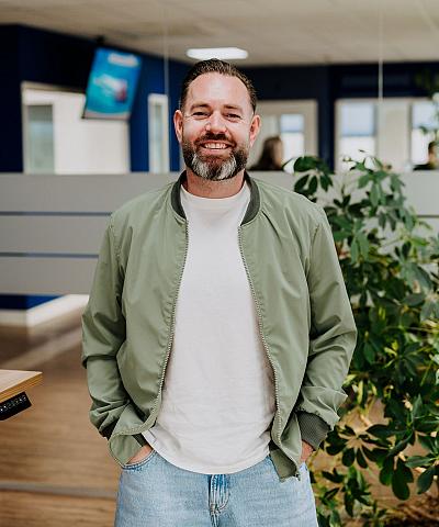 Medewerker zit lachend aan een bureau in een modern kantoor met computerschermen, blauw accent op de muur en een grote groene plant bij het raam.