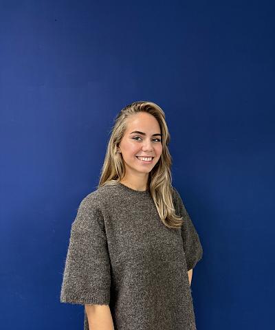 Vrouw met lang blond haar in een ruime, donkergrijze wollen trui staat tegen een effen donkerblauwe achtergrond, gefotografeerd vanaf ongeveer de heupen.