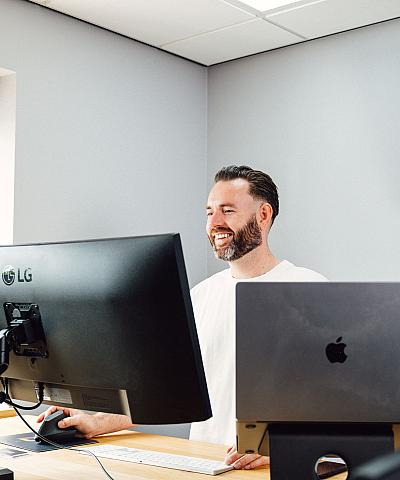 Man die lachend achter zijn bureau werkt met twee beeldschermen, in een modern kantoor met natuurlijk licht.