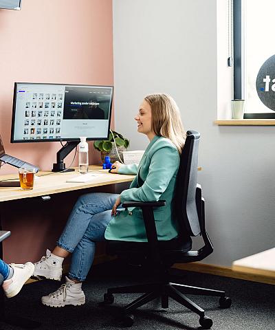 Vrouw die lachend aan het werk is achter haar bureau met laptop en beeldscherm, in een licht kantoor met roze muur en plant op de achtergrond.