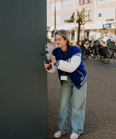 Jonge vrouw met blauwe jas filmt met een smartphone op een gimbal in een winkelstraat met fietsen en woonhuizen op de achtergrond.