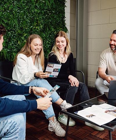 Vier collega’s die samen overleggen aan een lage tafel met laptop en tijdschriften, zittend in een ruimte met een groene plantenwand.