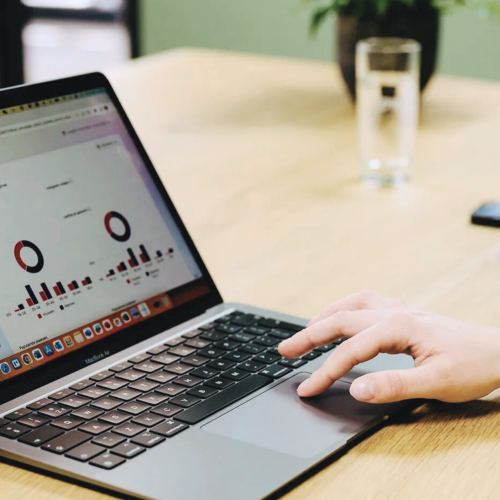 Laptop op houten tafel toont data en hand op toetsenbord klikt iets aan in kantooromgeving.