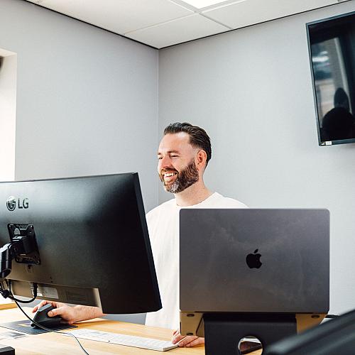 Een man werkt lachend achter twee schermen in een modern kantoor.