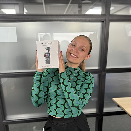 Vrouw glimlacht en houdt een doos met DJI Osmo Pocket 3 Creator Combo camera omhoog in een kantoorruimte.