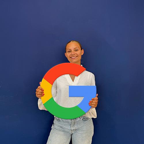 Vrouw die glimlachend een groot Google-logo vasthoudt, gekleed in een witte blouse en lichtblauwe jeans, voor een effen blauwe achtergrond.