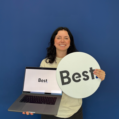 Een vrouw staat lachend voor een blauwe muur terwijl ze een laptop en een bord met het logo “Best.” vasthoudt.