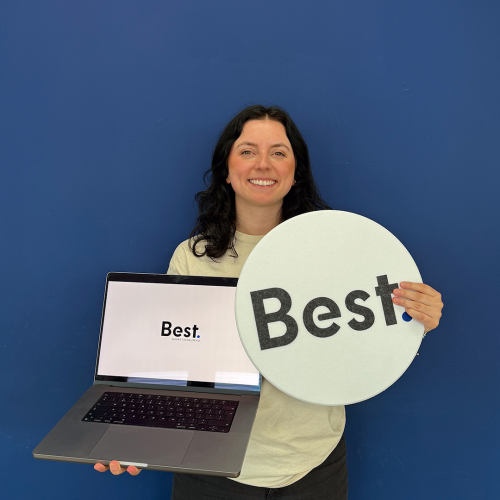 Een vrouw staat lachend voor een blauwe muur terwijl ze een laptop en een bord met het logo “Best.” vasthoudt.