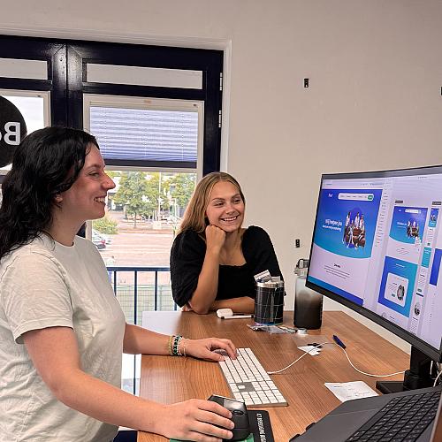 Twee collega’s werken samen achter een bureau terwijl ze een websiteontwerp op het computerscherm bekijken.