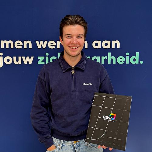 Jonge man met donkerblond haar en een blauwe trui poseert lachend voor een blauwe wand met de tekst “Samen werken aan jouw zekerheid.” Hij houdt een zwarte map met het PWA-logo vast.