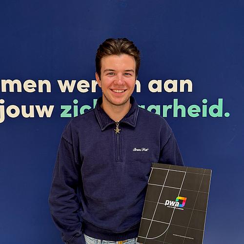 Jonge man met donkerblond haar en een blauwe trui poseert lachend voor een blauwe wand met de tekst “Samen werken aan jouw zekerheid.” Hij houdt een zwarte map met het PWA-logo vast.