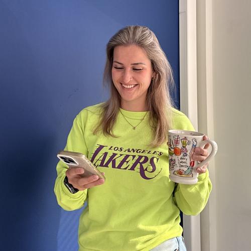 Vrouw die glimlachend op haar telefoon kijkt terwijl ze een mok vasthoudt, gekleed in een felgele Los Angeles Lakers-trui, voor een blauwe muur.