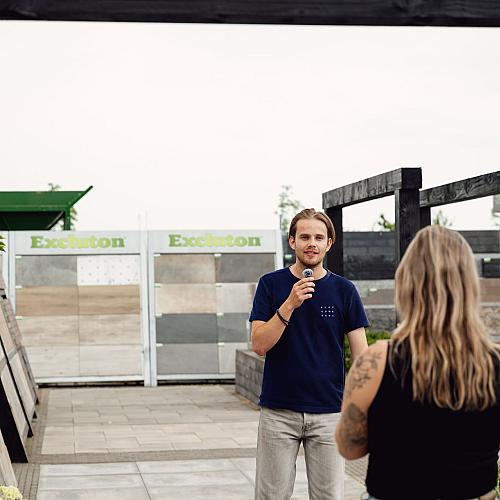 Man die buiten spreekt in een tegelshowtuin met een microfoon in de hand, terwijl een vrouw met lang blond haar naar hem luistert.