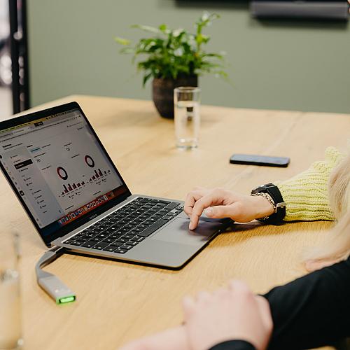 Twee mensen die samenwerken achter een laptop waarop grafieken en statistieken te zien zijn, aan een houten tafel met glazen water en planten.