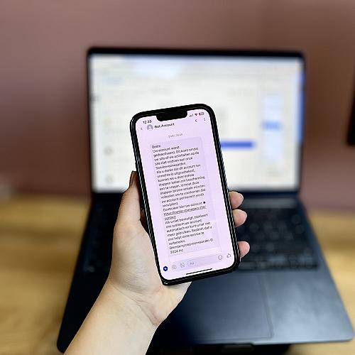 Hand die telefoon vasthoudt met daarop een bericht van Bot Account, op de achterkant geblurd een computer