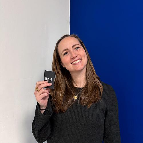 Vrouw met lang bruin haar houdt een zwart visitekaartje met het logo Best vast en glimlacht tegen een witte en blauwe achtergrond.