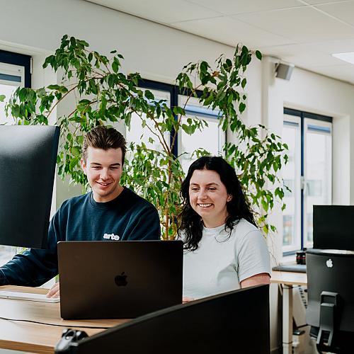 Twee collega’s werken samen achter een bureau met laptop en monitor in een licht kantoor met grote ramen en groene planten op de achtergrond.