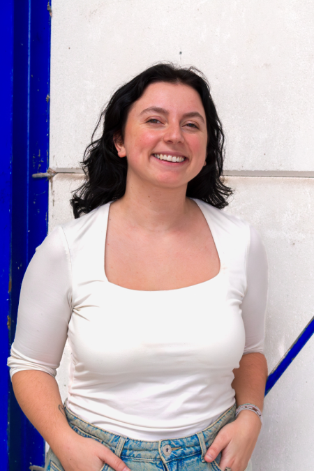 Vrouw met wit t-shirt en spijkerbroek leunt tegen blauw paal in loodsomgeving.