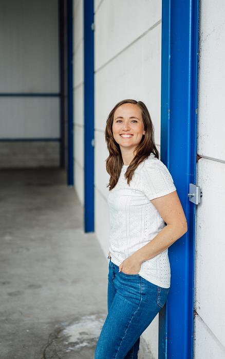 Vrouw met bruin haar leunt lachend tegen een witte muur met blauwe accenten in een industriële omgeving.